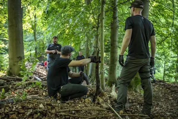 Zwei Personen arbeiten im Wald an einer Holzstruktur, während andere im Hintergrund stehen