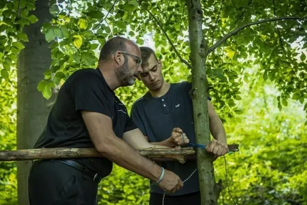 Zwei Personen befestigen eine Holzstange an einem Baum mit Schnüren