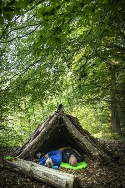 Zwei Personen liegen in einer Tarp-Notunterkunft aus Ästen im Wald