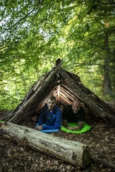 Zwei Personen sitzen in einer selbstgebauten Laubhütte im Wald
