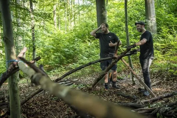 Zwei Personen stehen an einer selbstgebauten Seilbrücke aus Ästen im Wald