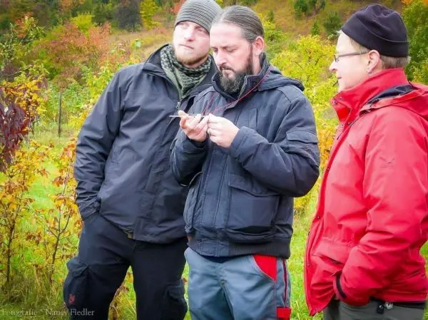 Drei Personen betrachten ein Gerät in der Natur zwischen buntem Laub