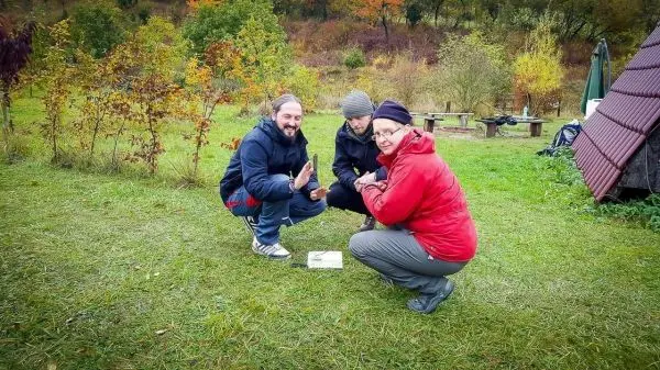 Drei Personen hocken auf dem Gras und betrachten ein Objekt auf dem Boden