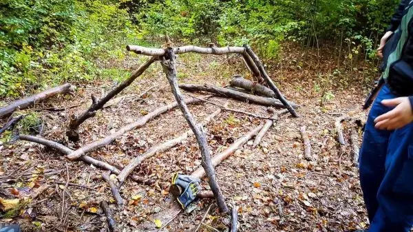Person steht neben einer selbstgebauten Unterkunft aus Ästen im Wald