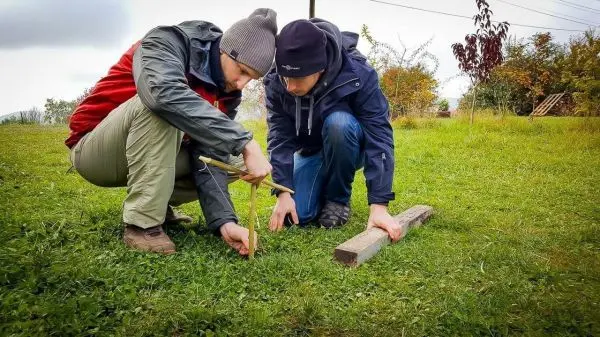 Zwei Personen konstruieren eine einfache Feuerbohrvorrichtung auf einer Wiese
