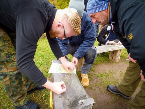 Karte wird mit einem Maßband von mehreren Personen auf einer Holzbank abgemessen