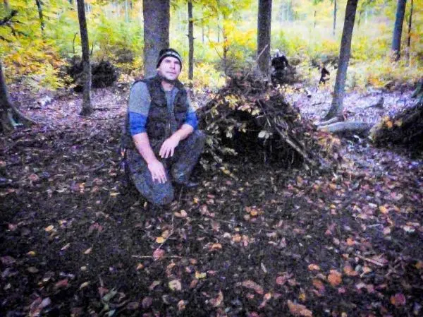 Mann kniet neben einer Laubhütte im Wald, umgeben von herbstlichen Blättern