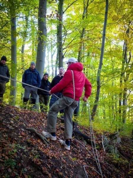 Person mit Seil über einen Abhang kletternd, umgeben von Bäumen und Zuschauern