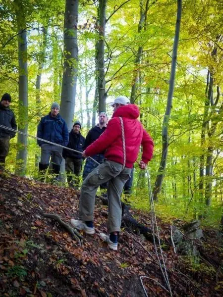 Person mit Seil über einen Abhang kletternd, umgeben von Bäumen und Zuschauern