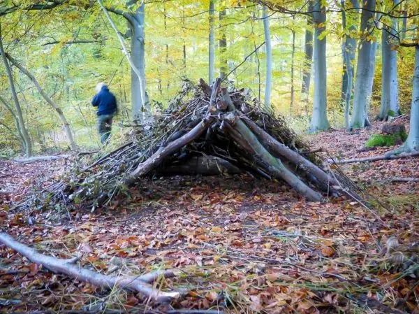 Person steht neben einer selbstgebauten Unterkunft aus Ästen und Blättern