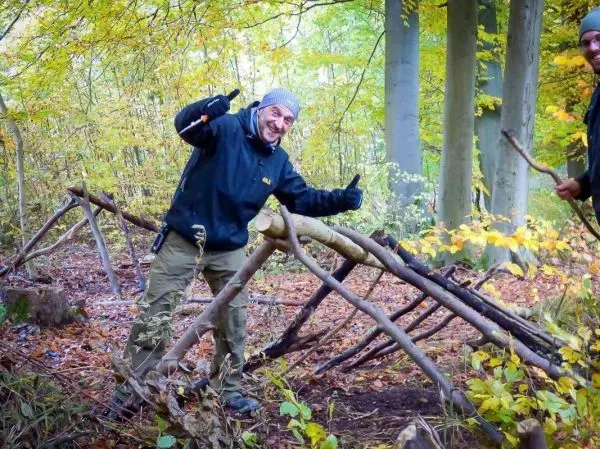 Person zeigt Daumen hoch neben selbstgebauter Struktur im Wald