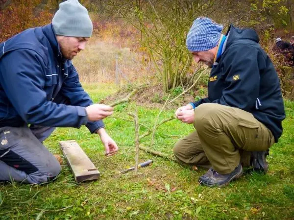 Zwei Personen binden mit Schnüren eine Holzstruktur aus Ästen auf dem Boden