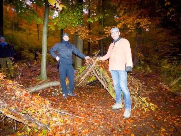 Zwei Personen stehen neben einer selbstgebauten Laubhütte im Wald