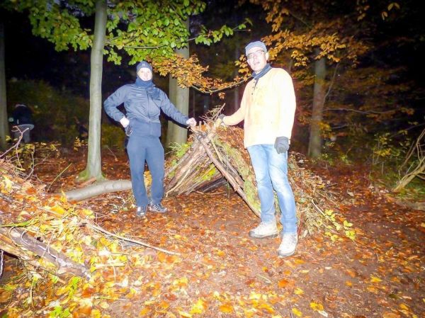 Zwei Personen stehen neben einer selbstgebauten Laubhütte im Wald