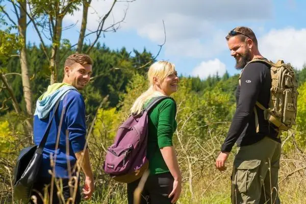 Drei Wandernde mit Rucksäcken lächeln sich an und gehen einen Waldweg entlang