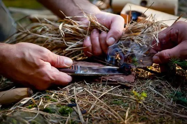 Hände halten Gras und ein Messer über Holzstücken beim Feuer machen