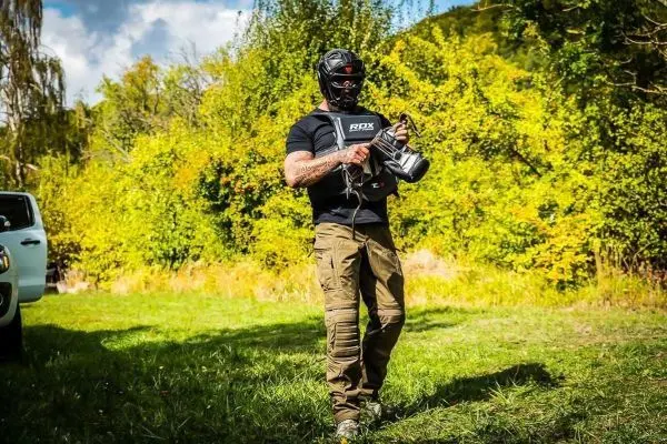 Person trägt eine Ausrüstung und geht über eine Wiese mit Bäumen im Hintergrund