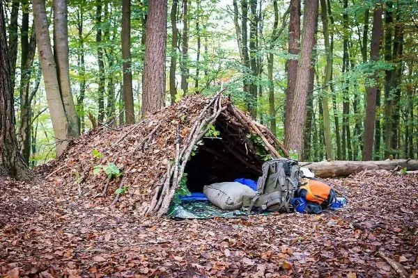 Tarp-Notunterkunft aus Ästen und Blättern mit Rucksack und Schlafsack davor