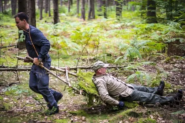 Zwei Personen ziehen eine improvisierte Trage aus Ästen durch den Wald