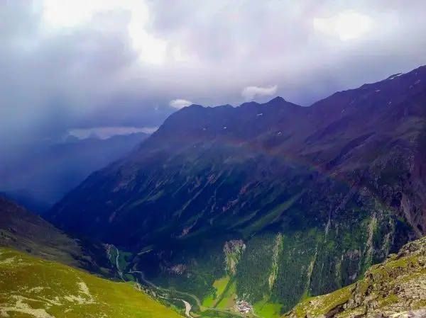 Berglandschaft mit Regenbogen über einem Tal und bewaldeten Hängen