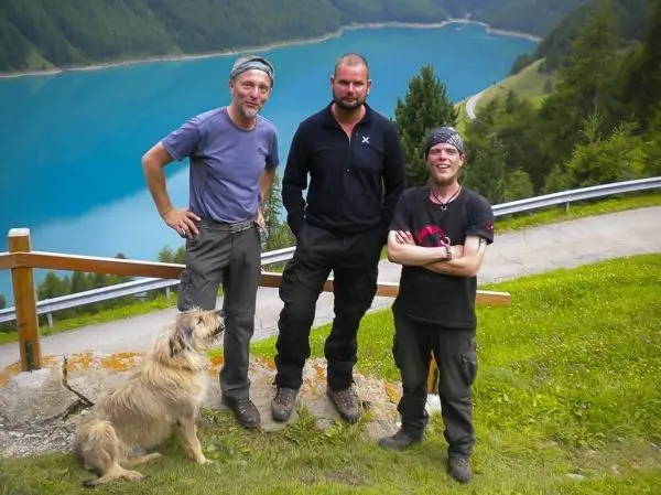 Drei Personen stehen auf einer Wiese mit einem Hund, im Hintergrund ein blauer See