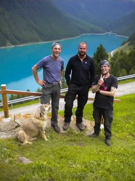 Drei Personen und ein Hund stehen auf einer Wiese mit Blick auf einen blauen See