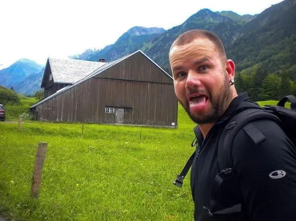 Ein Mann mit Rucksack steht vor einem Holzgebäude in einer grünen Landschaft