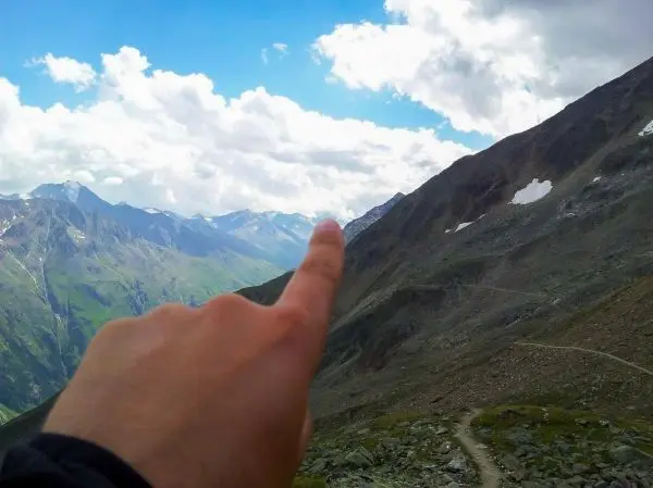 Hand zeigt auf einen Berg in einer gebirgigen Landschaft mit Wolken
