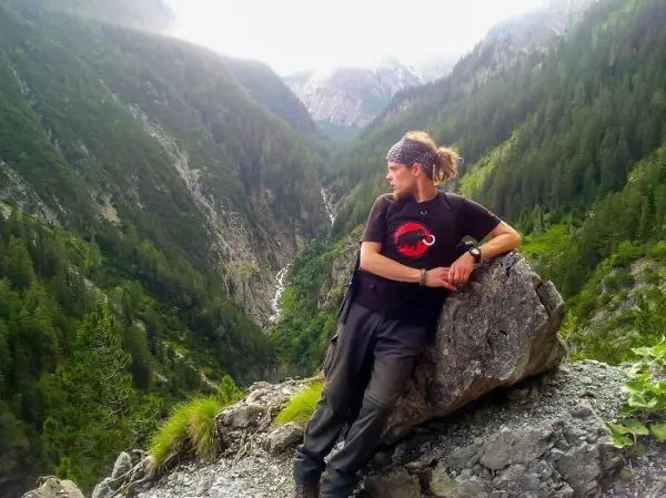 Mann lehnt an einem großen Stein und blickt ins Tal mit Bäumen und Bergen