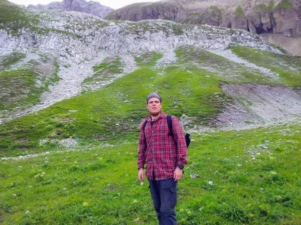 Mann mit Rucksack steht auf einer grünen Wiese vor einer felsigen Berglandschaft