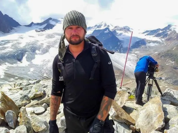 Mann steht auf einem Berggipfel, im Hintergrund schneebedeckte Berge und Kameraaufbau