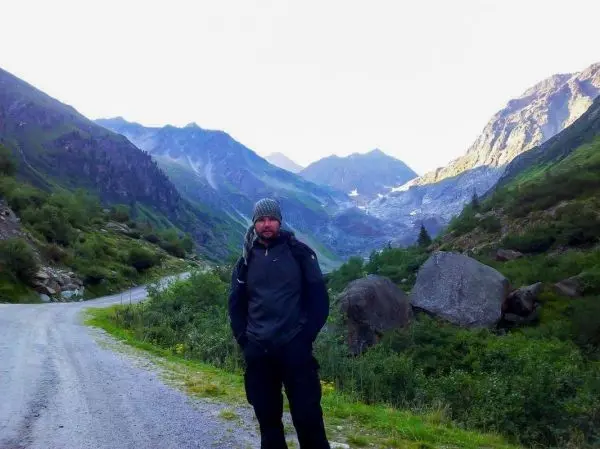 Mann steht auf einem Schotterweg in einer bergigen Landschaft mit Gletschern im Hintergrund