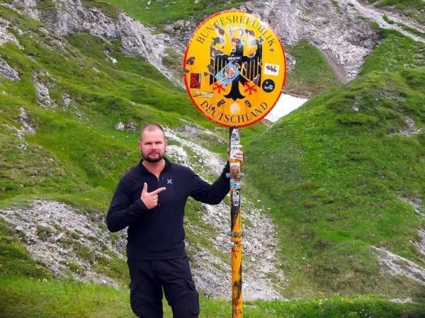 Mann zeigt auf ein Schild mit dem Schriftzug „Bundesrepublik Deutschland“ in einer Berglandschaft