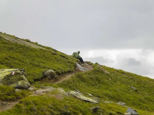 Person sitzt auf einem Hang neben einem Wanderweg, umgeben von Gras und Felsen