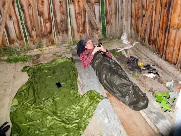 Schlafsack liegt auf Holzplanken, Person nutzt Smartphone in improvisierter Unterkunft