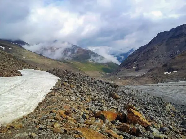 Schneebedeckte Fläche und steiniger Hang in einem Gebirgstal mit Wolken