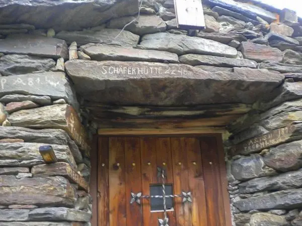 Steinmauer mit Holzverkleidung und Tür, Beschriftung über dem Eingang sichtbar