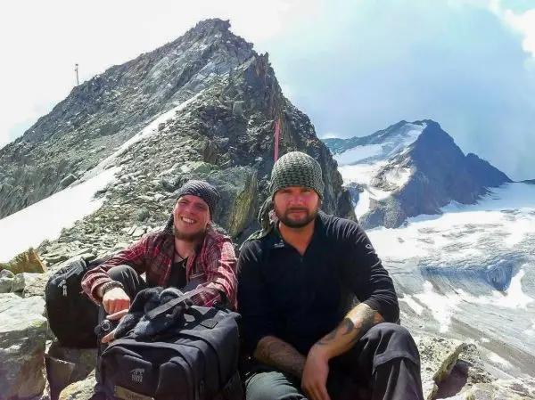 Zwei Personen sitzen auf einem Felsen mit Rucksäcken, im Hintergrund Berge und Gletscher