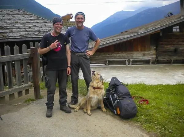 Zwei Wanderer stehen neben einem Hund und einem Rucksack vor einer Hütte
