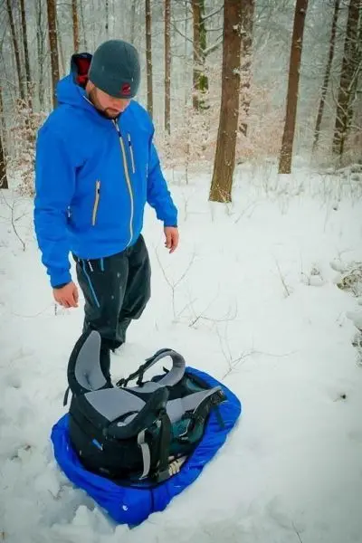Person in blauer Jacke steht im Schnee neben einem Rucksack
