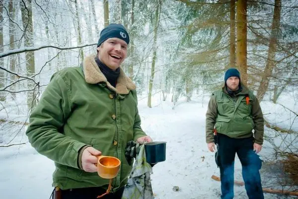 Zwei Personen im Schnee, eine hält Tassen, Bäume im Hintergrund