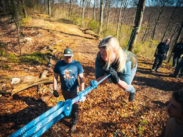 Frau überquert selbstgebaute Seilbrücke, während Männer sie sichern