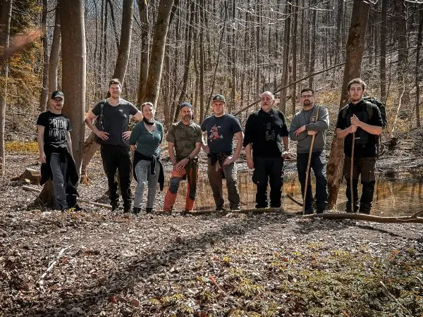 Gruppierung von acht Personen am Waldrand, einige halten Stöcke oder Werkzeuge