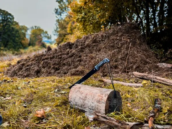 Holzstück mit Klappsäge auf dem Boden, im Hintergrund eine Erdhütte aus Laub