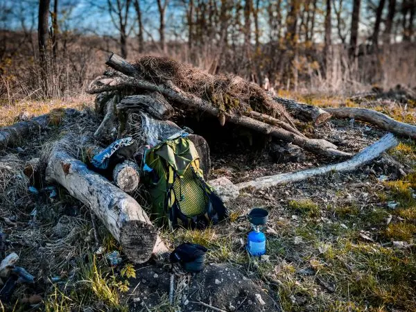 Tarp-Notunterkunft aus Ästen, Rucksack und Kocher auf dem Boden