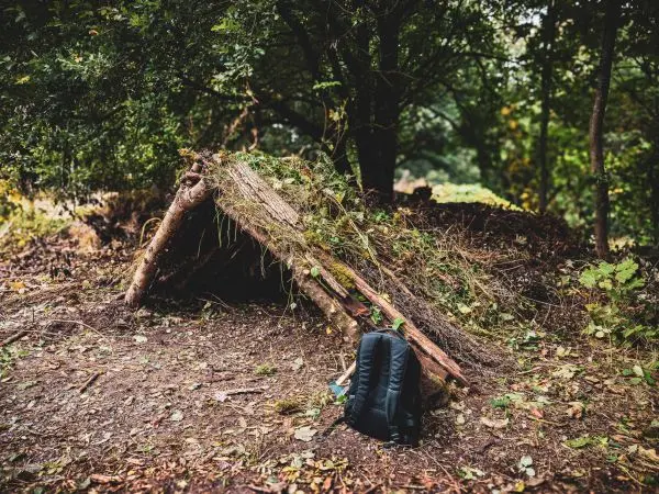 Tarp-Notunterkunft aus Ästen und Laub neben einem Rucksack im Wald