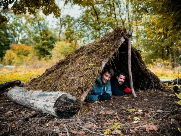 Zwei Personen sitzen in einer selbstgebauten Tarp-Notunterkunft aus Ästen und Laub
