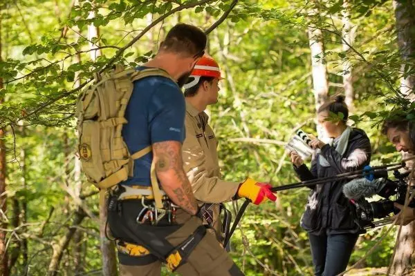 Drei Personen in Waldumgebung, eine Person hält Kamera, andere tragen Sicherheitsausrüstung
