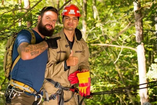 Zwei Personen in Kletterausrüstung stehen in einem Wald, eine Person zeigt Daumen hoch
