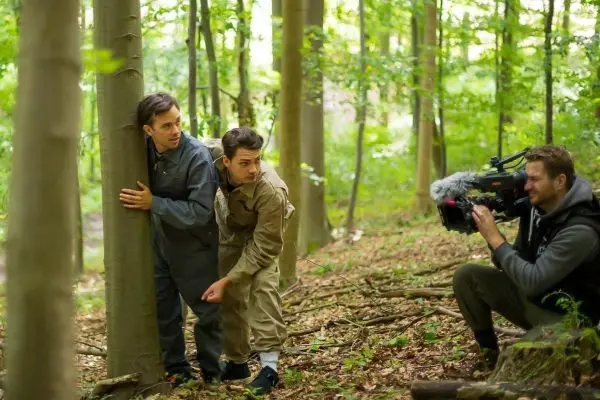 Zwei Personen lehnen an einem Baum, während ein Kameramann filmt